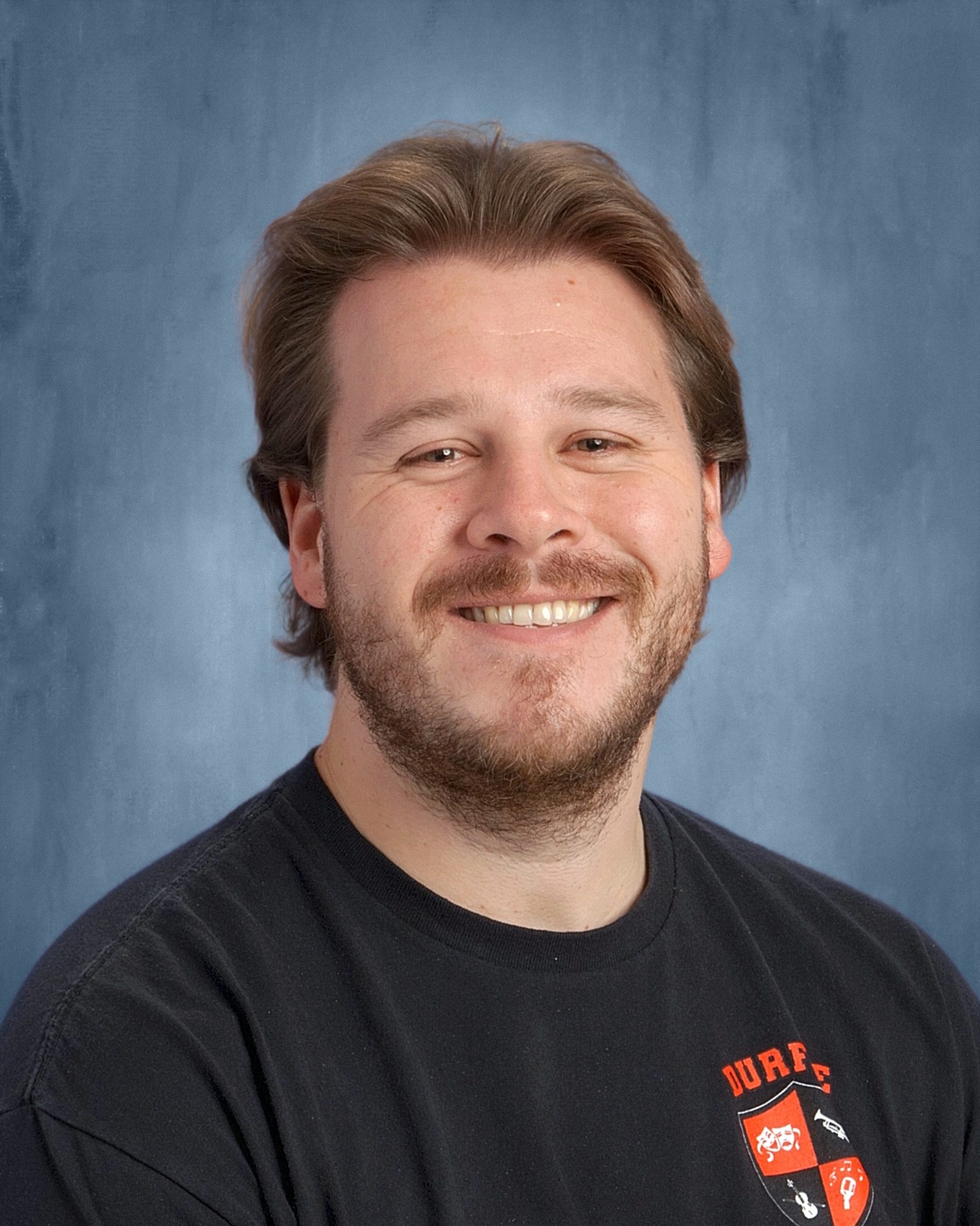 A white man with a short blonde beard wearing a black shirt partially showing the red text, "Durfee," and the Durfee Performing Arts symbol, looking directly at the camera with a calm expression.